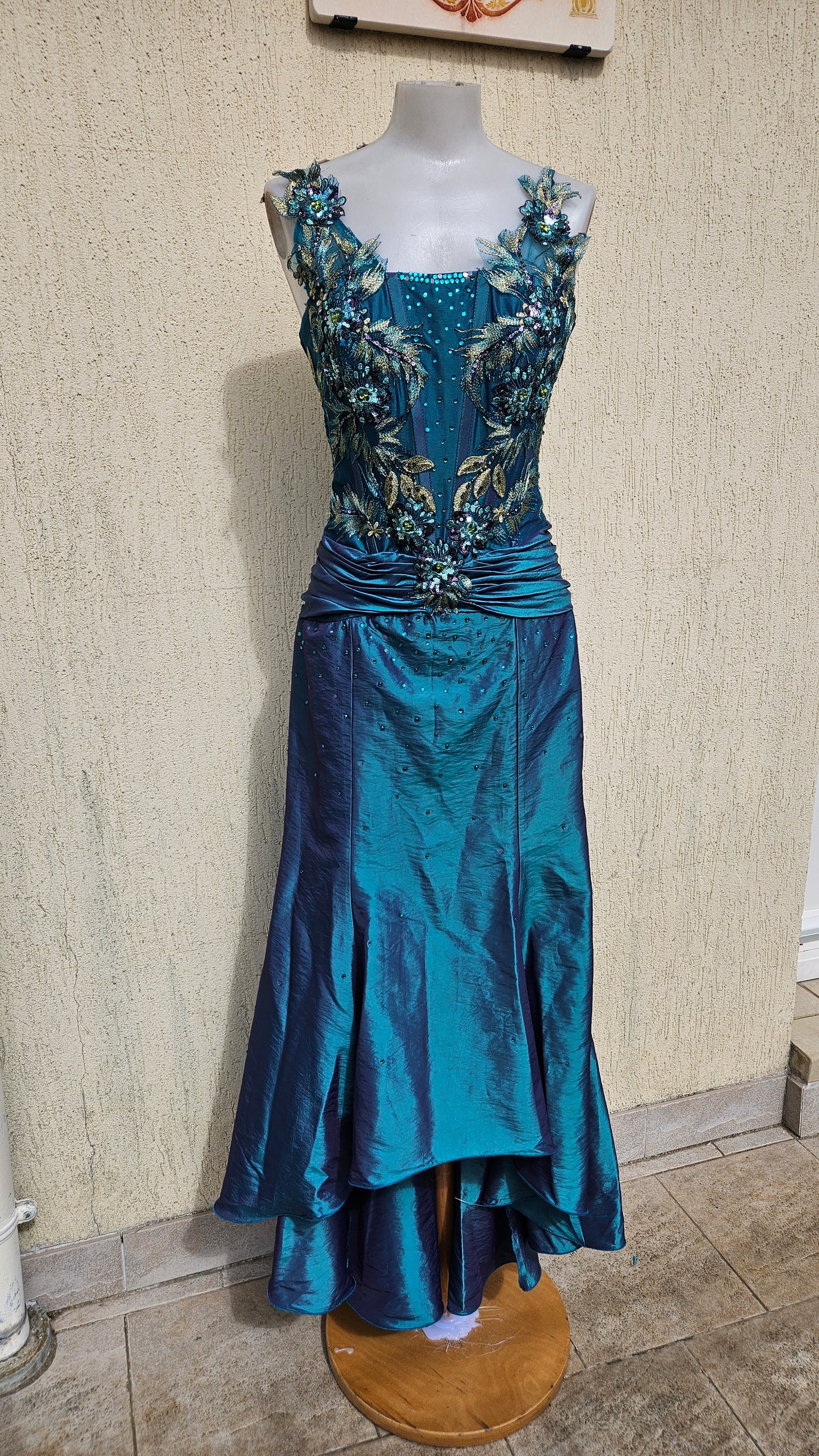Iridescent teal mermaid gown with floral 3D appliqués, gold embroidery, and sequin bodice. Features a ruched waist and layered high-low hem for formal events