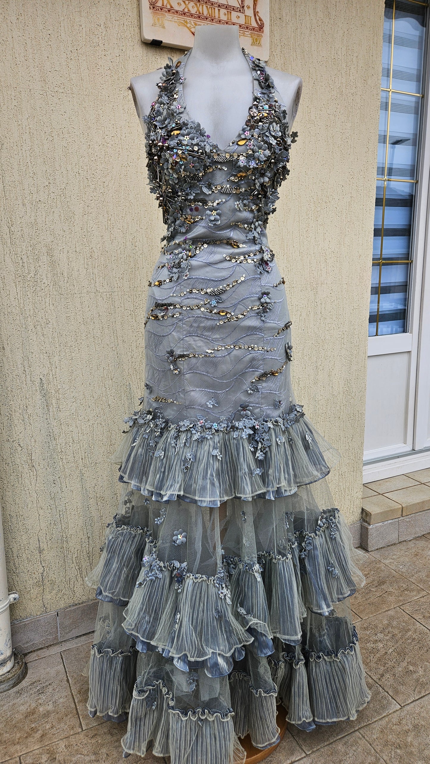 Silver-grey halter neck mermaid dress with 3D floral appliqués, gold sequins, and a tiered ruffled tulle skirt. Elegant vintage-style gown for formal events.